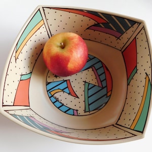 May include: A square ceramic bowl with a beige background and a colourful geometric design. The bowl has a red and yellow apple inside. The bowl's design includes black polka dots and colourful shapes in red, blue, and green.