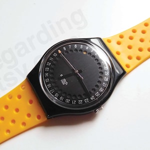 Peut inclure: Une montre-bracelet Swatch avec un cadran circulaire noir doté d'un anneau de date numéroté de 1 à 31, des aiguilles noires et un bracelet en silicone orange vif perforé. Le boîtier est en plastique noir avec une couronne argentée sur le côté.
