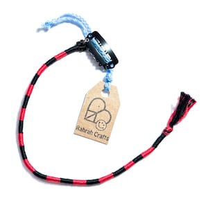 May include: A handmade bracelet with red and black thread, a black metal clip, and a light blue string. A small, brown tag with the text "Rahrah Crafts" is attached to the bracelet.