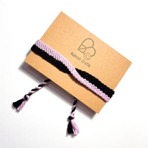 May include: A handmade friendship bracelet with black and light purple woven bands. The bracelet is displayed on a brown card with the text "Rahrah Crafts" and a logo. The bracelet has braided ends with black and light purple tassels.