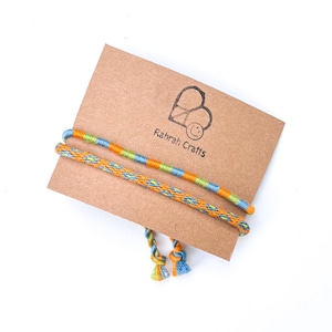 May include: Two handmade thread bracelets attached to a rectangular brown cardboard display card. One bracelet features alternating orange, light green, and blue segments. The other has a blue and orange diamond pattern. The card displays the text Rahrah Crafts.