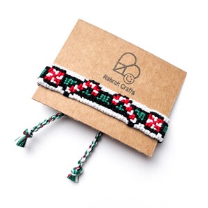 May include: A woven friendship bracelet in red, green, black, and white, displayed on a brown card. The bracelet features a detailed pattern and is finished with braided ties. The card has the text "Rahrah Crafts" and a logo.