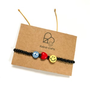 May include: A black beaded bracelet featuring a peace sign, a red heart, and a yellow smiley face charm. The bracelet is displayed on a brown card with the text "Rahrah Crafts".