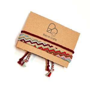 May include: Two handmade friendship bracelets with a chevron pattern in shades of red, green, brown, and pink. The bracelets are displayed on a brown card with the text "Rahrah Crafts" and a logo. The bracelets have braided ends.