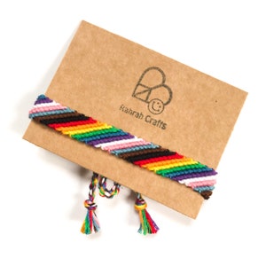 May include: A colorful friendship bracelet with a rainbow pattern, featuring a variety of colors including red, orange, yellow, green, blue, purple, pink, brown, black, and white. The bracelet is displayed on a brown card with the text "Rahrah Crafts".