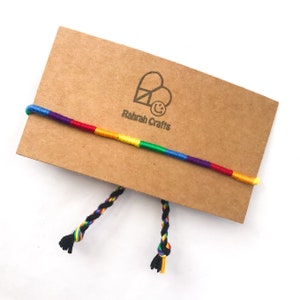 May include: Rainbow friendship bracelet with black string ties. The bracelet is on a brown card with the text "Rahrah Crafts" and a logo.