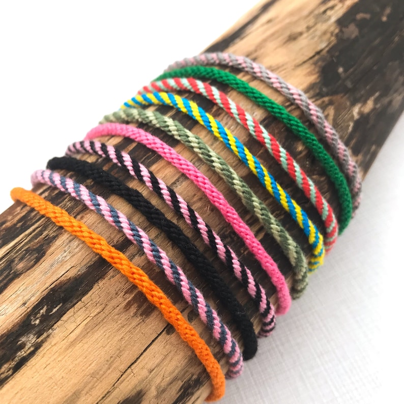 Puede incluir: Una colecci&oacute;n de pulseras de amistad coloridas hechas con cuerda trenzada. Las pulseras presentan varios patrones, incluyendo rayas, zigzags y colores s&oacute;lidos. Las pulseras est&aacute;n dispuestas en una pila sobre una superficie de madera.
