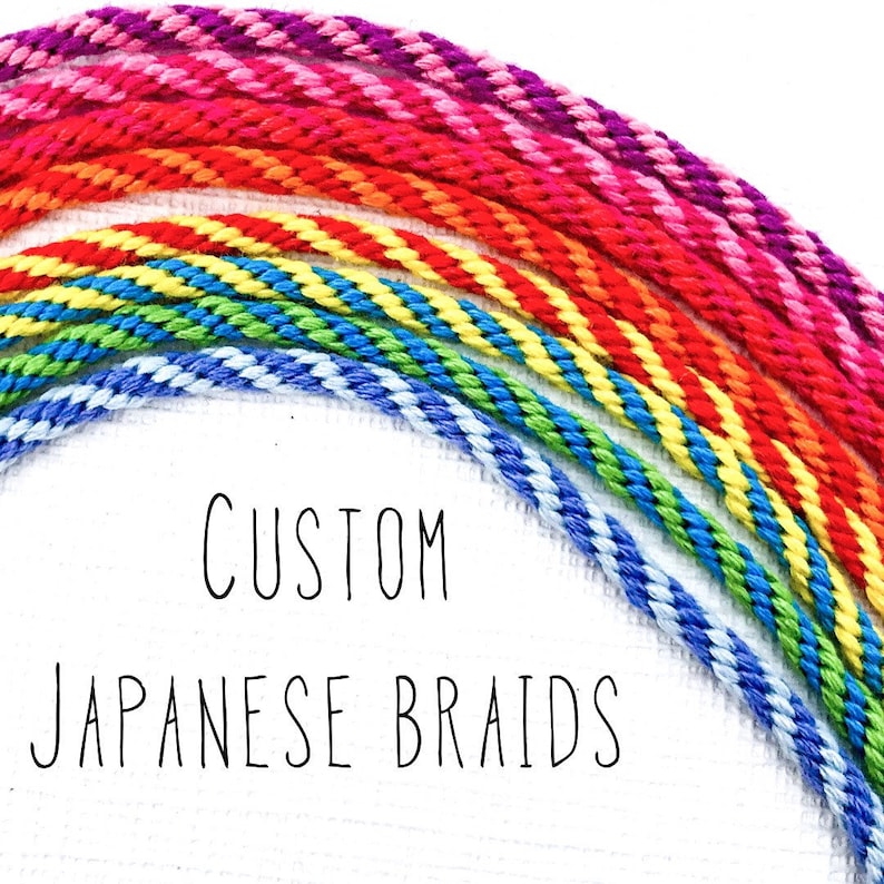 Puede incluir: Primer plano de cordones trenzados de colores dispuestos en forma de arco&iacute;ris. Los cordones est&aacute;n hechos de un material suave y texturizado y son perfectos para hacer pulseras u otras joyas. El texto "CUSTOM JAPANESE BRAIDS" est&aacute; escrito debajo de los cordones.