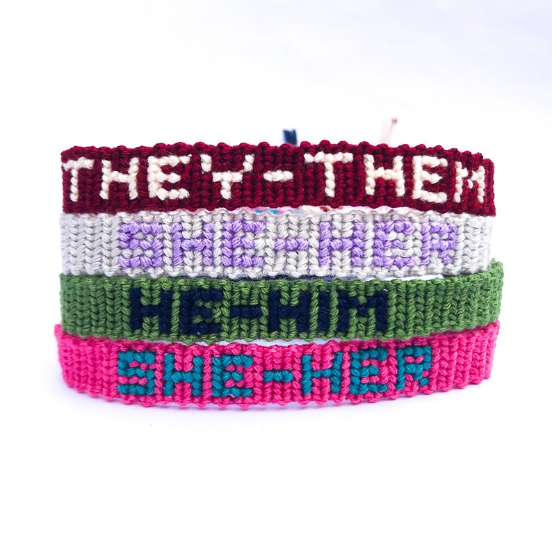 K&ouml;nnte beinhalten: Vier Freundschaftsarmb&auml;nder in verschiedenen Farben mit den W&ouml;rtern "She-Her", "He-Him", "They-Them" und "She-Her", die in den Stoff eingewebt sind.