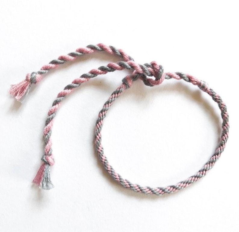 Puede incluir: Un brazalete de amistad trenzado de color rosa y gris con cierre anudado.