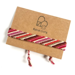 May include: A colourful friendship bracelet with red, pink, white, brown and orange stripes. The bracelet is on a brown card with the text "Rahrah Crafts" printed on it.