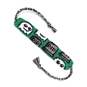 May include: A woven friendship bracelet featuring a green background with a repeating pixelated ghost and block pattern in white, black, and gray. The bracelet has braided ties at both ends, incorporating green, black, and white thread colors.
