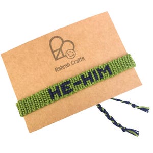 K&ouml;nnte beinhalten: Gr&uuml;nes Freundschaftsarmband mit den Worten "He + Him", die in marineblauem Garn gewebt sind. Das Armband wird auf einer braunen Karte mit einem Herz und den Worten "Rahrah Crafts" pr&auml;sentiert.