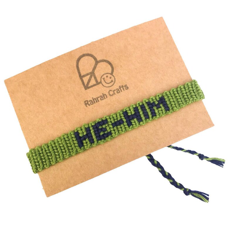 K&ouml;nnte beinhalten: Gr&uuml;nes Freundschaftsarmband mit den Worten "He + Him", die in marineblauem Garn gewebt sind. Das Armband wird auf einer braunen Karte mit einem Herz und den Worten "Rahrah Crafts" pr&auml;sentiert.