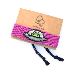May include: A pink friendship bracelet with a grey and green alien spaceship design. The bracelet is on a brown card with the text "Rahrah Crafts".