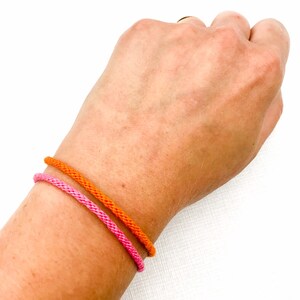 Puede incluir: Dos pulseras en una mu&ntilde;eca. La pulsera superior es naranja y la pulsera inferior es rosa.