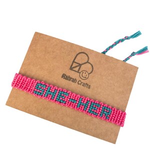 K&ouml;nnte beinhalten: Pinkes und t&uuml;rkisfarbenes Freundschaftsarmband mit dem Wort "SHE-HER", das in das Design eingewebt ist. Das Armband wird auf einer braunen Karte mit dem Text "Ratrah Crafts" und einem Friedenssymbol gezeigt.
