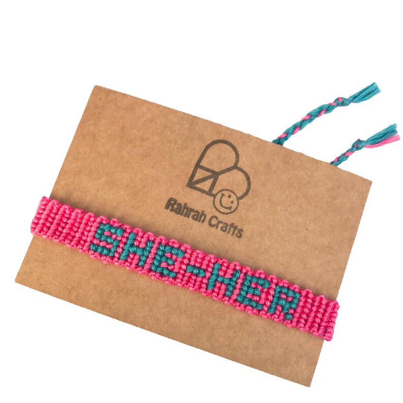 K&ouml;nnte beinhalten: Pinkes und t&uuml;rkisfarbenes Freundschaftsarmband mit dem Wort "SHE-HER", das in das Design eingewebt ist. Das Armband wird auf einer braunen Karte mit dem Text "Ratrah Crafts" und einem Friedenssymbol gezeigt.