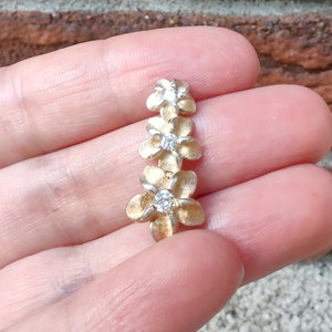 Peut inclure: Un charme en forme de fleur en or avec trois pierres de zircone cubique blanches. Le charme est en forme de trois fleurs de frangipanier.