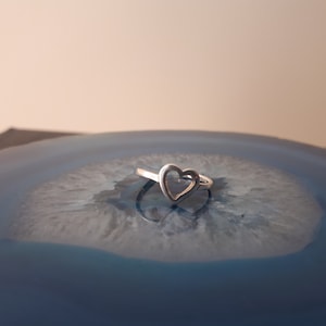 May include: A silver ring with a heart-shaped cutout design. The ring is sitting on a blue and white agate surface.