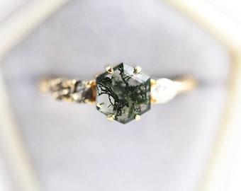 May include: A gold ring featuring a central hexagonal moss agate stone with dark green inclusions. The band is accented with small, textured gold details and a pear-shaped white stone on one side, set in a six-prong gold setting.