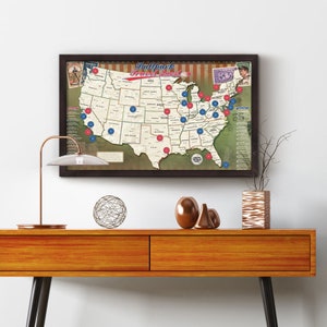 May include: A framed map of the United States with red and blue dots marking locations. The map is titled "Ballpark Travel Quest" and features a baseball theme. The map is hanging on a white wall above a wooden table with decorative objects.