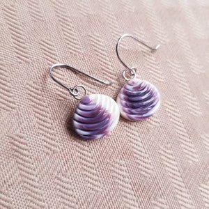 May include: A pair of seashell earrings with sterling silver ear wires. The earrings have a round, shell-shaped design with alternating bands of white and purple. The earrings are displayed on a textured, light pink surface.