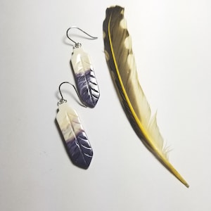May include: A pair of white and purple feather-shaped earrings with silver hooks. The earrings are displayed next to a brown and yellow feather.