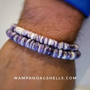 May include: A double-strand bracelet made of purple and white shell beads. The bracelet is worn on a person's wrist.