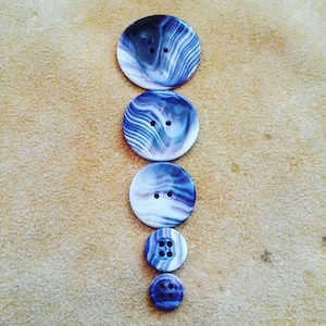 May include: A set of five iridescent buttons with a swirling blue and white pattern. The buttons are arranged in a vertical line, with the largest button at the top and the smallest button at the bottom.