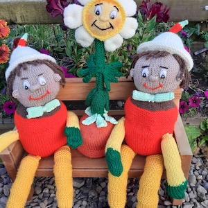 Puede incluir: Dos personajes de juguete de punto, que se asemejan a personas, están sentados en un banco de madera. Llevan ropa roja y amarilla con detalles verdes. Una flor de punto con una cara sonriente está detrás de ellos.