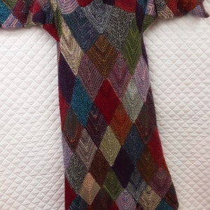 K&ouml;nnte beinhalten: Ein langer, bunter Strickpullover mit einem Rautenmuster. Der Pullover ist aus verschiedenen Farben gefertigt, darunter Rot, Blau, Gr&uuml;n und Lila.