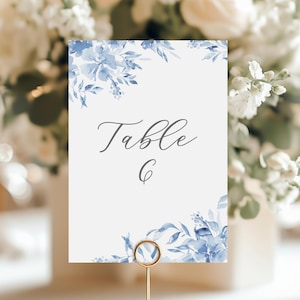May include: A white table number card with blue watercolour floral designs. The card is on a gold metal stand and reads "Table 6".