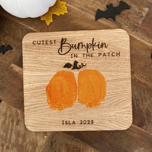 May include: A square wooden plaque featuring the engraved text CUTEST BUMPKIN IN THE PATCH and ISLA 2023. The centre displays two orange handprints or footprints arranged to resemble a pumpkin, complete with an engraved stem and leaf detail.
