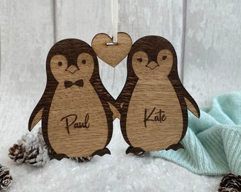 Könnte beinhalten: Holz-Pinguin-Paar-Anhänger mit einem Herz in der Mitte. Die Pinguine halten sich an den Händen und haben die Namen "Paul" und "Kate" eingraviert.