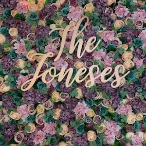 May include: A floral wall backdrop featuring a wooden sign that reads "The Joneses". The wall is adorned with a mix of flowers, including roses, hydrangeas, and foliage.