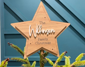 May include: A wooden star tree topper with the text "The Wilkinson Family Christmas" in brown lettering. The star is a light brown color and has a white outline.