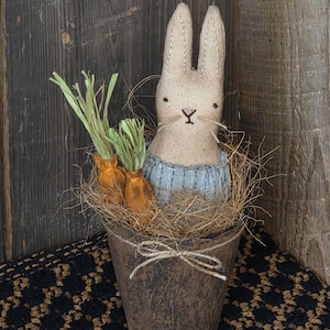 May include: A handcrafted bunny doll with a light beige fabric body, black eyes, and a stitched mouth. It sits in a rustic brown pot filled with straw and two orange carrot decorations. The pot is tied with twine, creating a charming Easter decoration.