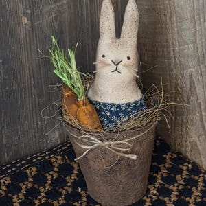 May include: A rustic Easter bunny doll with a carrot in a decorative pot. The bunny has a beige burlap body, a blue floral patterned dress, and stitched facial features. The pot is brown and tied with twine, sitting on a black and brown patterned surface.