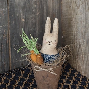 May include: A handcrafted Easter bunny doll with a burlap body and long ears, wearing a blue floral print shirt. It sits in a brown pot with two orange carrot decorations and green stems. The pot is tied with twine.