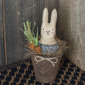May include: A rustic Easter bunny decoration. The bunny is made of burlap and has a stitched face. It sits in a brown pot with faux carrots and greenery. The pot is tied with a twine bow. The background is a wooden wall.