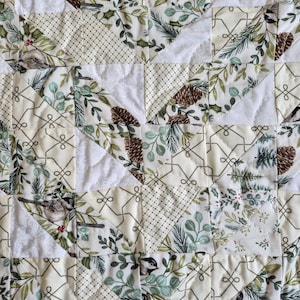 May include: A quilted fabric featuring a patchwork design of cream, white, and green. The pattern includes illustrations of pinecones, evergreen branches, small birds, and geometric lines. The fabric has a visible stitched texture throughout.