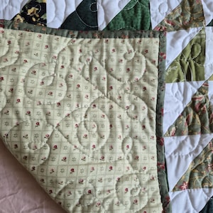 May include: A quilted blanket featuring a patchwork design of white and various green fabric triangles. The underside displays a cream-colored fabric with a small red floral print and a grid pattern, finished with a dark green floral border.