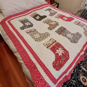 May include: A handmade Christmas quilt featuring a repeating pattern of festive stockings in various colors and patterns. The quilt has a white background with a red border and is placed on a bed with white sheets.