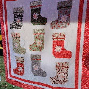 May include: A handmade Christmas quilt featuring nine decorative stockings in various patterns and colors, including red, green, and black. Each stocking has a star-shaped design in the center. The quilt is bordered with a red and white snowflake pattern.
