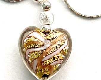 Puede incluir: Un collar de cadena de plata con un colgante en forma de corazón. El colgante está hecho de vidrio transparente con remolinos dorados y rosados en su interior. El colgante está unido a la cadena con una cuenta de plata.