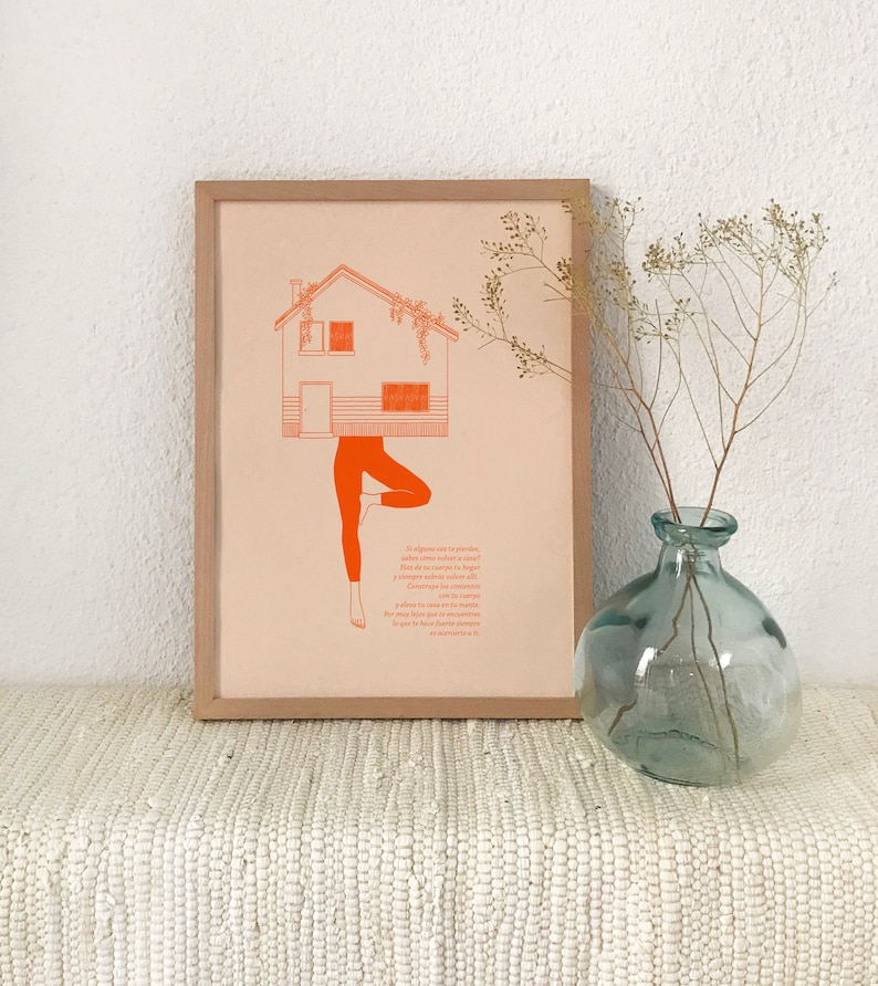May include: A framed print with a peach background featuring a house with a green roof and a person doing a yoga pose. The house is upside down and the person's legs are the house's foundation. The print includes a Spanish language quote about finding your place in the world.