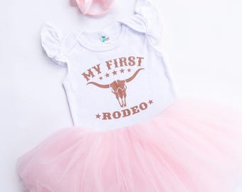 May include: A white baby bodysuit with the text "MY FIRST RODEO" in brown, a pink tutu skirt, and a matching pink bow. The bodysuit features a steer skull graphic. The outfit is designed for a baby.
