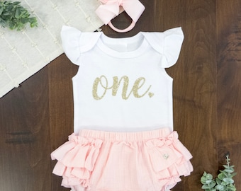 May include: A white onesie with gold glitter lettering that says "one" and a pink ruffled diaper cover. A pink bow headband is included.