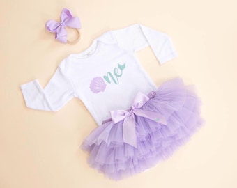 May include: A white long-sleeved bodysuit with a mermaid-themed design, a lavender tutu skirt, and a matching bow headband. The bodysuit features the word "one" and a seashell graphic. The tutu has a large bow.